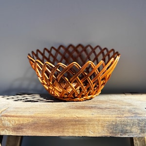 Puede incluir: Una cesta tejida con un patrón de diamantes. La cesta es de mimbre marrón claro y está sobre una superficie de madera. El diseño abierto y el color cálido de la cesta sugieren un uso decorativo o de almacenamiento.