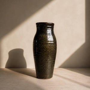 Könnte beinhalten: Eine hohe, dunkelgrüne Keramikvase mit strukturierter Oberfläche. Die Vase hat einen leicht ausgestellten Rand und einen abgerundeten Körper. Die Vase wird auf einer neutralen Oberfläche mit einem Schatten an der Wand dahinter präsentiert.