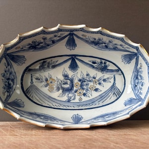 May include: An oval ceramic serving dish with two handles, featuring a white base with blue floral patterns and swags. The rim and handle edges are accented with gold trim. The central design depicts a cluster of blue roses and small yellow flowers.