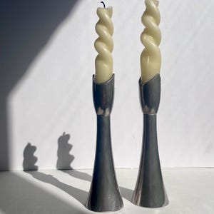 Puede incluir: Dos candelabros plateados, cada uno con una vela retorcida de color crema. Los candelabros tienen una base acampanada y una parte superior curvada. El fondo es blanco con una sombra de las velas.