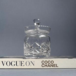 Peut inclure: Un pot en verre transparent avec un motif en cristal et un couvercle. Le pot est posé sur un livre blanc avec le texte "VOGUE ON COCO CHANEL" imprimé sur la couverture.