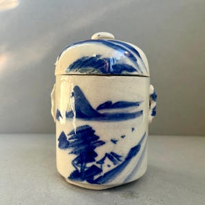 Puede incluir: Un pequeño tarro de cerámica con tapa, con un diseño de paisaje azul y blanco. El tarro tiene una tapa redondeada con un pequeño pomo y dos pequeñas asas. El diseño del paisaje incluye montañas y árboles.