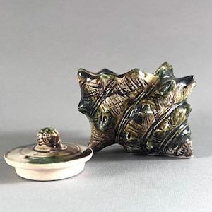 May include: A ceramic seashell-shaped container with a matching lid. The container is mainly green and brown with a textured surface. The lid is off-white with green and brown accents. The container is likely for decorative or storage purposes.