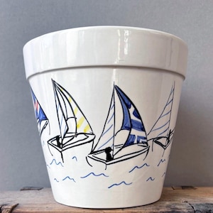 Puede incluir: Una maceta de cerámica blanca con una ilustración pintada a mano de varios veleros sobre el agua. El diseño incluye contornos negros de barcos con detalles en azul, amarillo y rojo en las velas sobre un fondo de color blanco.