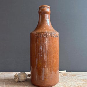 May include: A brown glazed stoneware bottle with a cylindrical body and a narrow neck. The word SCHWEPPES is embossed in capital letters on the upper portion of the bottle. The surface has a glossy finish with subtle variations in the brown tone.