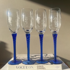 May include: Four clear champagne flutes with etched grape designs. Each glass has a vibrant blue twisted stem and base. The glasses are displayed on a book with the text "VOGUE ON COCO CHANEL".