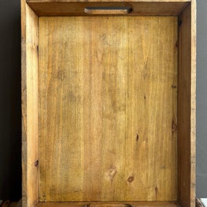 May include: A rectangular wooden tray with a natural light brown finish, featuring visible wood grain and knots. The tray has high vertical sides and two oval-shaped cutout handles, one centred on the top edge and one on the bottom edge.