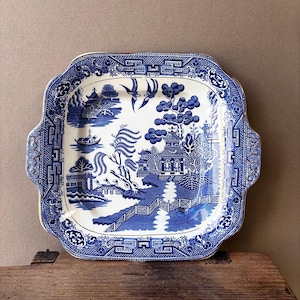 May include: A blue and white ceramic serving platter with a classic Willow pattern design. The square platter features a detailed landscape scene with bridges, trees, and birds. The edges have a decorative border and small handles.