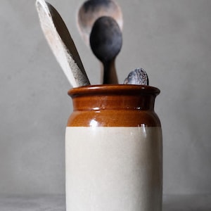 Puede incluir: Un tarro cilíndrico de cerámica con un diseño bicolor que presenta un borde esmaltado en marrón brillante y una base de color crema mate. Varias cucharas de madera de distintos tamaños y tonos marrones se encuentran en su interior.
