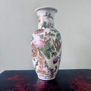 Może przedstawiać: Biały ceramiczny wazon z kwiatowym wzorem. Wazon ma smukły, wydłużony kształt z szeroką podstawą i lekko rozszerzoną krawędzią. Kwiatowy wzór obejmuje różowe, brązowe i zielone kwiaty i liście. Wazon stoi na ciemnej drewnianej powierzchni.