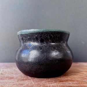 May include: A small, rounded ceramic pot with a dark, textured black glaze that transitions into a lighter, mottled finish. The rim features a distinct band of muted teal-green glaze, creating a contrast against the deep, dark body of the vessel.