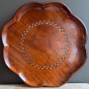May include: A round wooden tray with a scalloped edge and a smooth, polished finish. The centre of the tray features a circular inlay pattern composed of alternating light and dark wood triangles arranged in a repeating geometric border design.