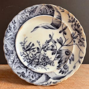 Pode incluir: Um prato de cerâmica redondo com borda festonada, apresentando um padrão de transferência azul escuro sobre um fundo creme. O design inclui um pássaro pousado num ramo, motivos florais e bordas geométricas com craquelado visível.