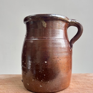 May include: A brown glazed ceramic pitcher with a cylindrical body, a thick rounded rim, and a single curved handle on the side. The surface features a glossy finish with visible variations in colour, small speckles, and a light-coloured chip on the rim.