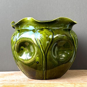 Peut inclure: Un cache-pot en céramique vert olive foncé brillant avec un bord large et ondulé. L'extérieur présente un motif en relief composé de grandes indentations arrondies et de crêtes verticales en forme de feuilles sur le corps.