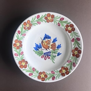 Könnte beinhalten: Ein weißer Keramikteller mit handbemaltem Design eines Vogels mit blauen und orangefarbenen Flügeln, umgeben von einem Rand aus orangefarbenen Blumen und grünen Blättern.