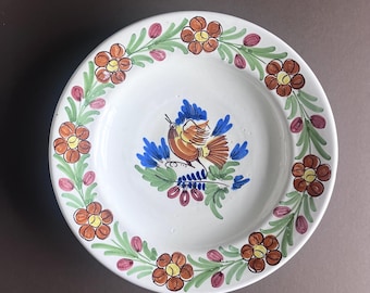 Pode incluir: Um prato de cerâmica branco com um desenho pintado à mão de um pássaro com asas azuis e laranja, rodeado por uma borda de flores laranja e folhas verdes.