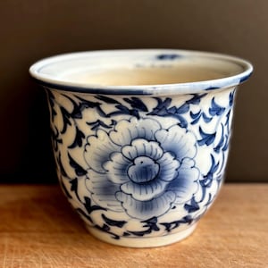 Puede incluir: Una maceta de cerámica con base blanca y un patrón floral azul pintado a mano. El diseño muestra una flor central grande rodeada de intrincadas enredaderas. El borde está rematado con una fina línea azul y el interior es de color crema sólido.