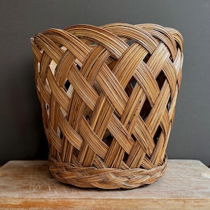 May include: A round, open-weave basket constructed from thin, light and dark brown natural reeds. The design features a wide, crisscross lattice pattern throughout the body, finished with a tightly braided circular base and a reinforced rim.