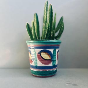 May include: A small, decorative ceramic pot with a green, blue, and red painted design, holding a green cactus plant. The pot has a cylindrical shape with horizontal stripes and abstract shapes. The cactus has multiple upright stems.