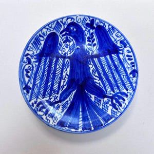 Op de afbeelding: Een ronde, handbeschilderde keramische plaat met een blauw adelaarsontwerp. De adelaar is gecentreerd op de plaat, met uitgespreide vleugels. De rand van de plaat heeft een decoratieve rand van blauwe bloemen en geometrische patronen. De plaat is wit met blauwe accenten.