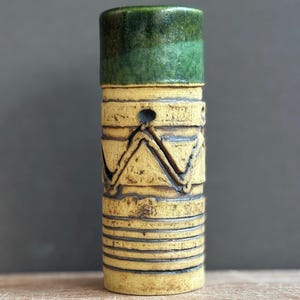 May include: A cylindrical ceramic vase featuring a smooth, glossy dark green glazed top section. The lower portion is finished in a matte, textured beige glaze with incised horizontal lines and a prominent zigzag pattern encircling the body.
