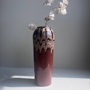 May include: A tall, cylindrical vase with a maroon base and a decorative top featuring a wavy pattern in black, beige, and brown. The vase holds a delicate arrangement of dried, silvery-white seed pods, adding a natural element to the design.