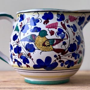 May include: A white ceramic jug featuring a hand-painted design of a yellow bird with green and red accents, surrounded by blue flowers and green foliage. The jug has a curved handle, a wide spout, and horizontal yellow and black lines.
