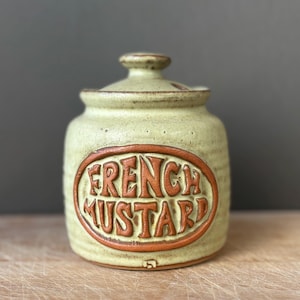 May include: A light green ceramic mustard jar with a matching lid. The jar features a prominent oval-shaped brown embossed label on the front with the text "FRENCH MUSTARD" in raised lettering. The surface has a speckled, textured finish.