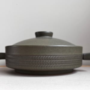 May include: A covered, olive-green ceramic casserole dish. The dish has a rounded lid with a small knob and two rectangular handles. The body of the dish features a textured, repeating chevron pattern around its circumference. The dish is designed for cooking and serving.