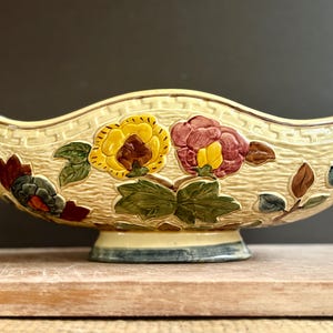 Pode incluir: Uma taça de cerâmica cor de creme com rebordo recortado e superfície texturizada com um padrão de grega. O exterior é decorado com motivos florais em relevo pintados à mão em amarelo, rosa, verde e castanho, sobre uma base de pedestal.