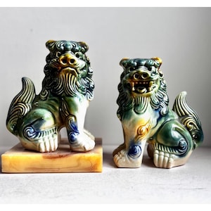 May include: Two ceramic guardian lion statues. The statues are green, yellow, and white with detailed features. They sit on rectangular bases. The lions have a fierce expression and are a traditional Asian decorative item.