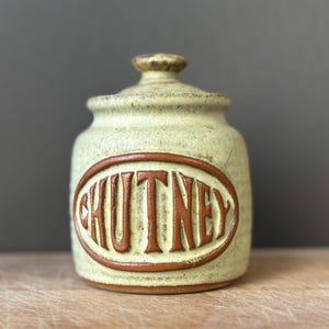 May include: A cylindrical ceramic jar with a speckled light green glaze and a matching lid. The front features a raised, dark brown oval frame containing the embossed text CHUTNEY in bold, capital letters, creating a textured appearance.