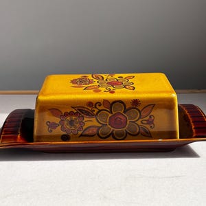 May include: A rectangular ceramic butter dish featuring a mustard yellow lid with a crackled glaze finish and a dark brown base. The lid is decorated with a floral pattern in shades of brown, orange, and red, with matching floral motifs on the front.