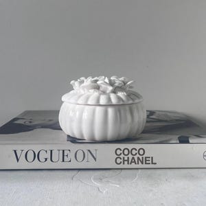 May include: A white ceramic trinket box with a floral lid rests on a book with the words "VOGUE ON COCO CHANEL" printed on the cover. The box has a ribbed design and is a soft, off-white colour.