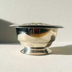 May include: A small, silver-coloured bowl with a wide, flat rim and a decorative pattern on the lid. The bowl has a pedestal base and a polished, reflective surface. The item is likely a decorative piece or a serving dish.