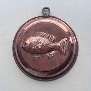 May include: A circular copper mould featuring a detailed embossed fish design. The fish is centrally positioned within the mould, and a small loop is attached at the top for hanging. The mould has a polished finish.