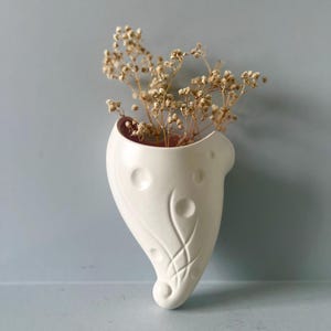Puede incluir: Un jarrón de pared de cerámica blanca con forma de lágrima, que contiene flores secas. El jarrón tiene una superficie texturizada con elementos decorativos circulares y lineales. El fondo es azul grisáceo claro.