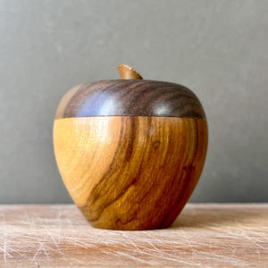 May include: A handcrafted wooden apple-shaped container featuring a two-tone design. The upper lid is made of dark brown wood, while the lower body displays a lighter, golden-brown wood grain with natural swirls and a small wooden stem on top.