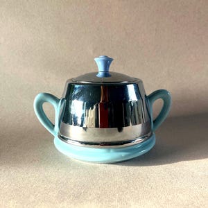 May include: A vintage sugar bowl with a chrome lid and a light blue base. The lid has a small, light blue knob on top.