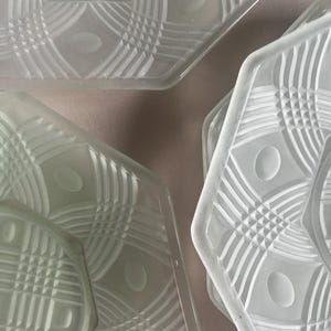 May include: Close-up of several clear glass plates with a geometric design. The plates have a hexagonal shape with etched patterns of lines, ovals, and diamond shapes. The glass has a frosted appearance.