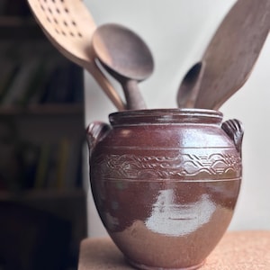 May include: A brown ceramic jar with two handles and a decorative band near the top, containing wooden kitchen utensils. The utensils include a slotted spoon, a spatula, and a round spoon.