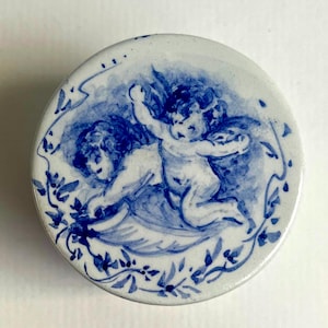 May include: Round, white ceramic trinket dish with a hand-painted design in shades of blue. The design features two cherubs with wings, surrounded by a floral border. The cherubs are depicted in a playful pose.