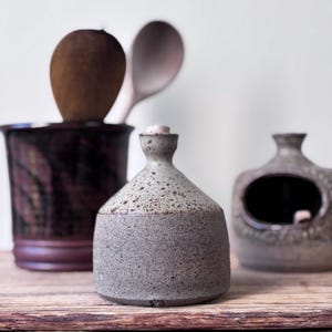 May include: A selection of handmade ceramic kitchen items. A speckled grey bottle with a cork stopper is in the foreground. Behind it is a dark brown utensil holder with wooden spoons. A small, square container is to the right.