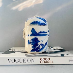 May include: A small, white ceramic jar with a lid, decorated with blue brushstrokes showing a landscape. The jar has two small handles and is on a Vogue On Coco Chanel book.