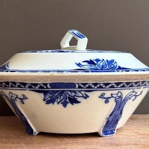 May include: A white ceramic serving bowl with a lid, featuring a blue floral and geometric pattern. The bowl has a wide rim, a tapered base with small feet, and a central handle on the lid. The design includes intricate blue leaf motifs.