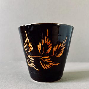 Peut inclure: Une tasse en céramique noire avec un motif de feuilles dorées. La tasse a une forme évasée et un intérieur blanc. Le motif de feuilles est gravé sur la surface, créant un effet texturé. Idéale pour les boissons.