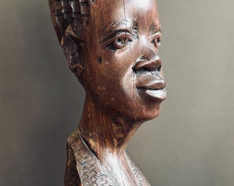 May include: A hand-carved wooden bust of a person with a textured hairstyle, defined facial features, and a V-neck garment. The sculpture features a dark brown finish with visible wood grain and intricate carved patterns on the clothing and hair.