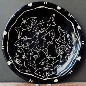 May include: A round ceramic plate with a glossy black glaze featuring a hand-drawn white line art pattern of multiple fish swimming in various directions. The rim of the plate is decorated with a series of white dots and short parallel lines.