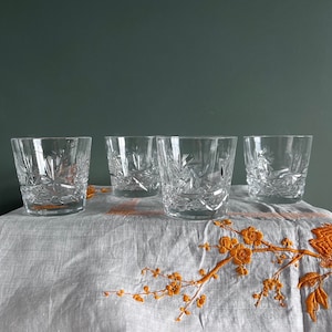 May include: Four clear, cut-glass tumblers with starburst and geometric patterns. The glasses are arranged on a white cloth with orange floral embroidery. The tumblers are designed for serving beverages.